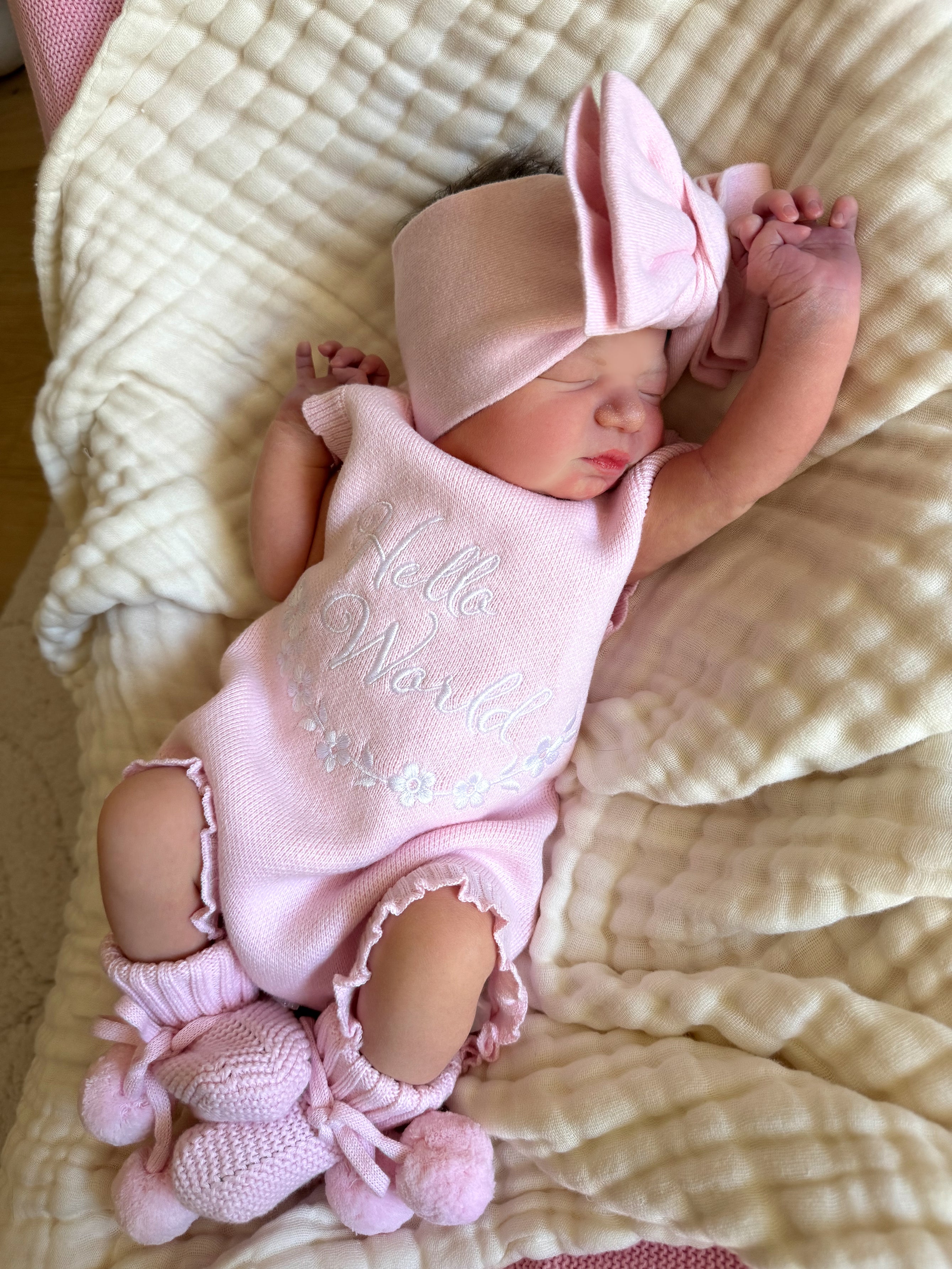 our pretty pink hello world has scalloped edges on short sleeve for arms and delicate knitted frill on legs. At the crotch and down the centre and on shoulder we have added pink resin flower buttons. Matches with our bonnet booties beanie and other pretty pink items. Hello world type in the centre is white surrounded by delicate flowers. The type is in running writing with capital letters at the start - beautiful for baby girl 