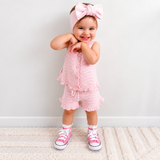 A toddler girl wearing a pink knit romper with ruffle trim, a matching pink bow headband, and pink sneakers, standing on a cream rug against a light gray wall.