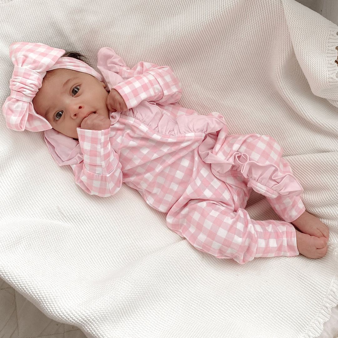 baby dressed in pink gingham frilly onesie with matching one size bow laying on white blanket