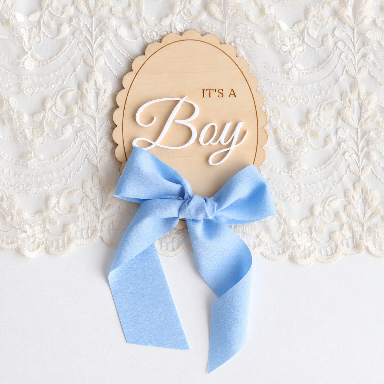 A wooden baby shower sign that reads 'IT'S A Boy' in white script, tied with a light blue satin bow, displayed on a white lace background.