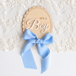 A wooden baby shower sign that reads 'IT'S A Boy' in white script, tied with a light blue satin bow, displayed on a white lace background.
