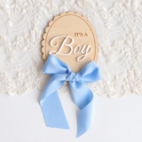 A wooden baby shower sign that reads 'IT'S A Boy' in white script, tied with a light blue satin bow, displayed on a white lace background.