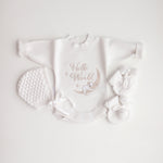 A flat lay of a white baby romper with 'Hello World' text and a koala illustration, accompanied by a bonnet and booties on a white background.