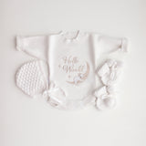 A flat lay of a white baby romper with 'Hello World' text and a koala illustration, accompanied by a bonnet and booties on a white background.