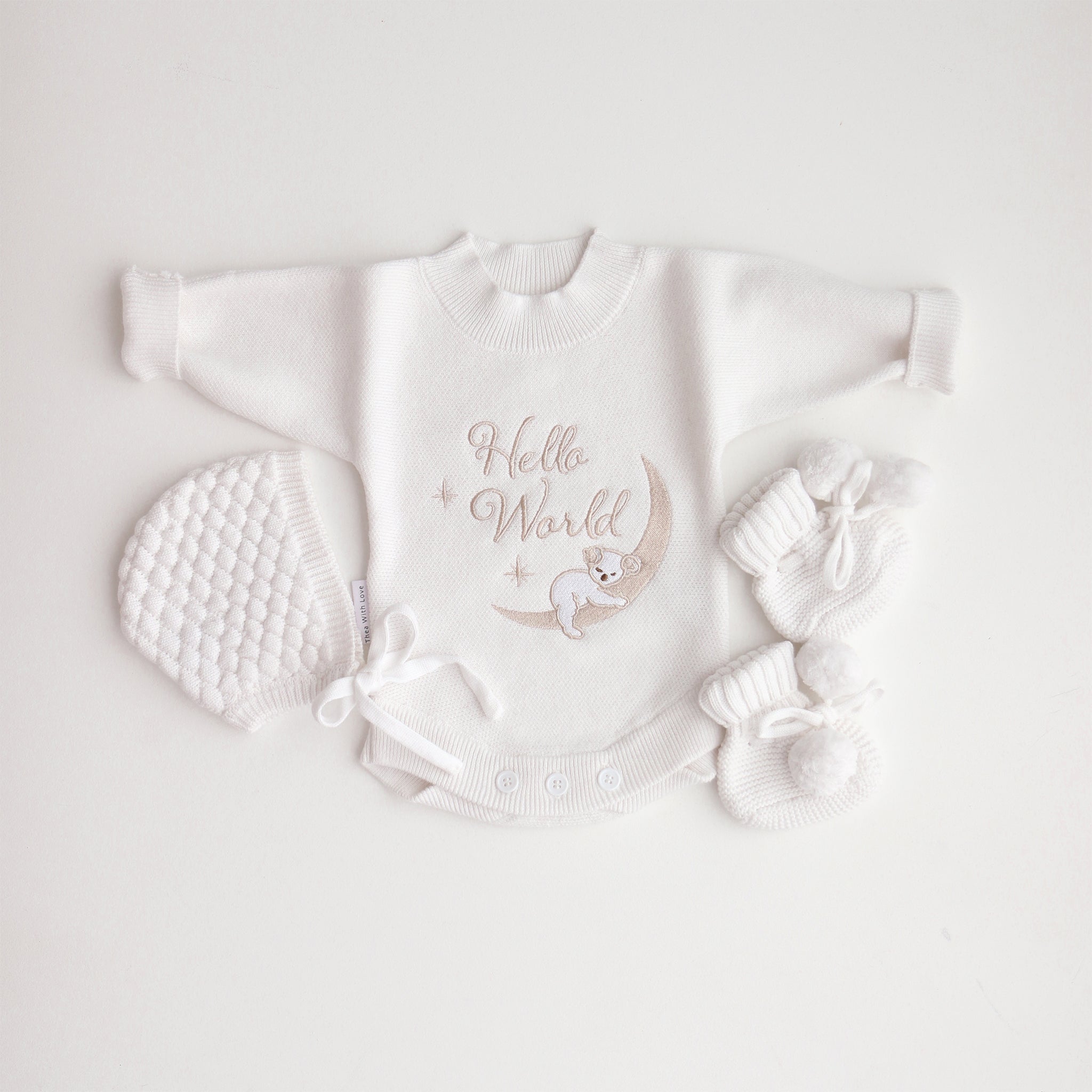 A flat lay of a white baby romper with 'Hello World' text and a koala illustration, accompanied by a bonnet and booties on a white background.