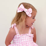 Young girl wearing a pink checkered dress with a large bow in her hair against a plain background