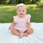 Baby in a pink checkered outfit sitting on a white blanket with a grassy background