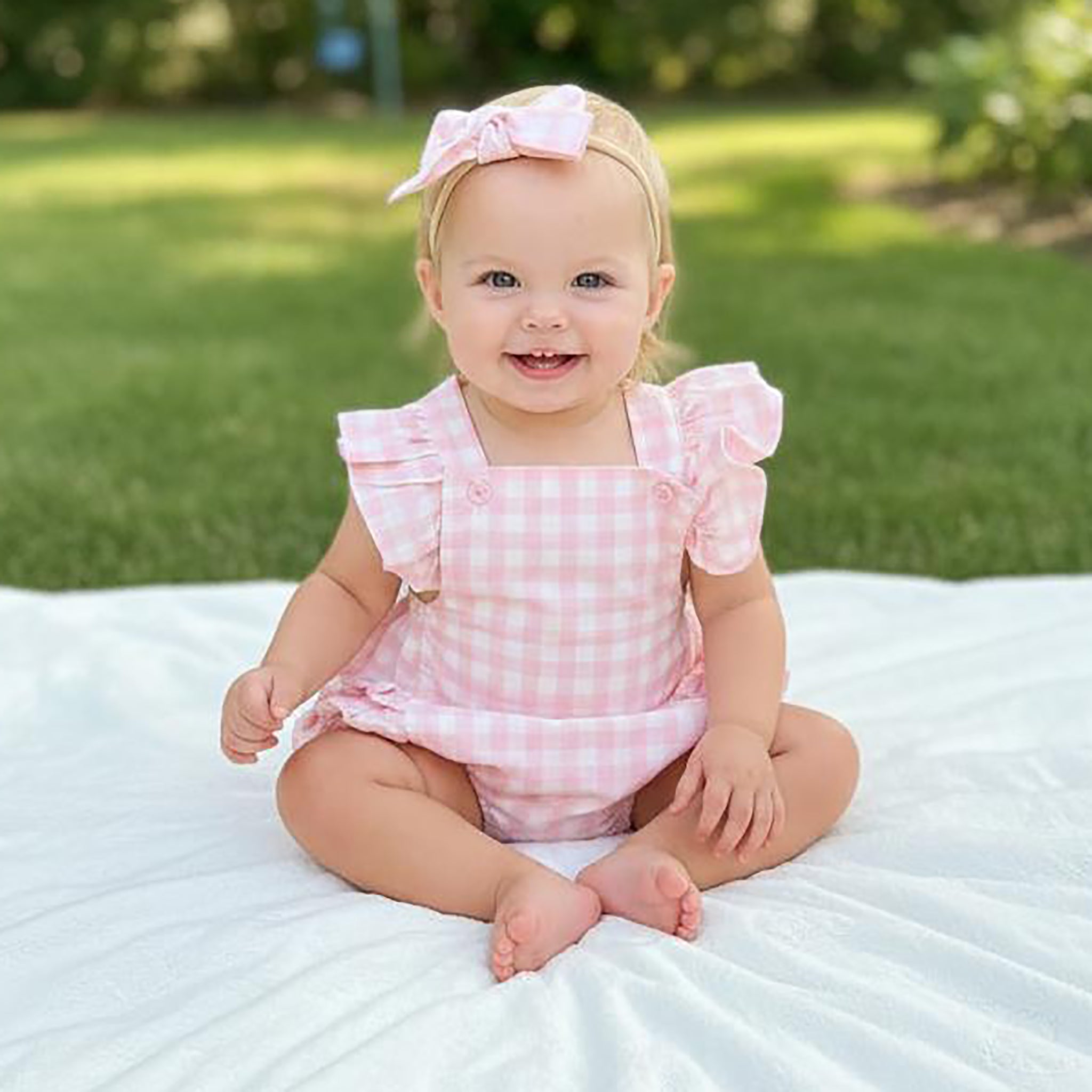 Baby in a pink checkered outfit sitting on a white blanket with a grassy background