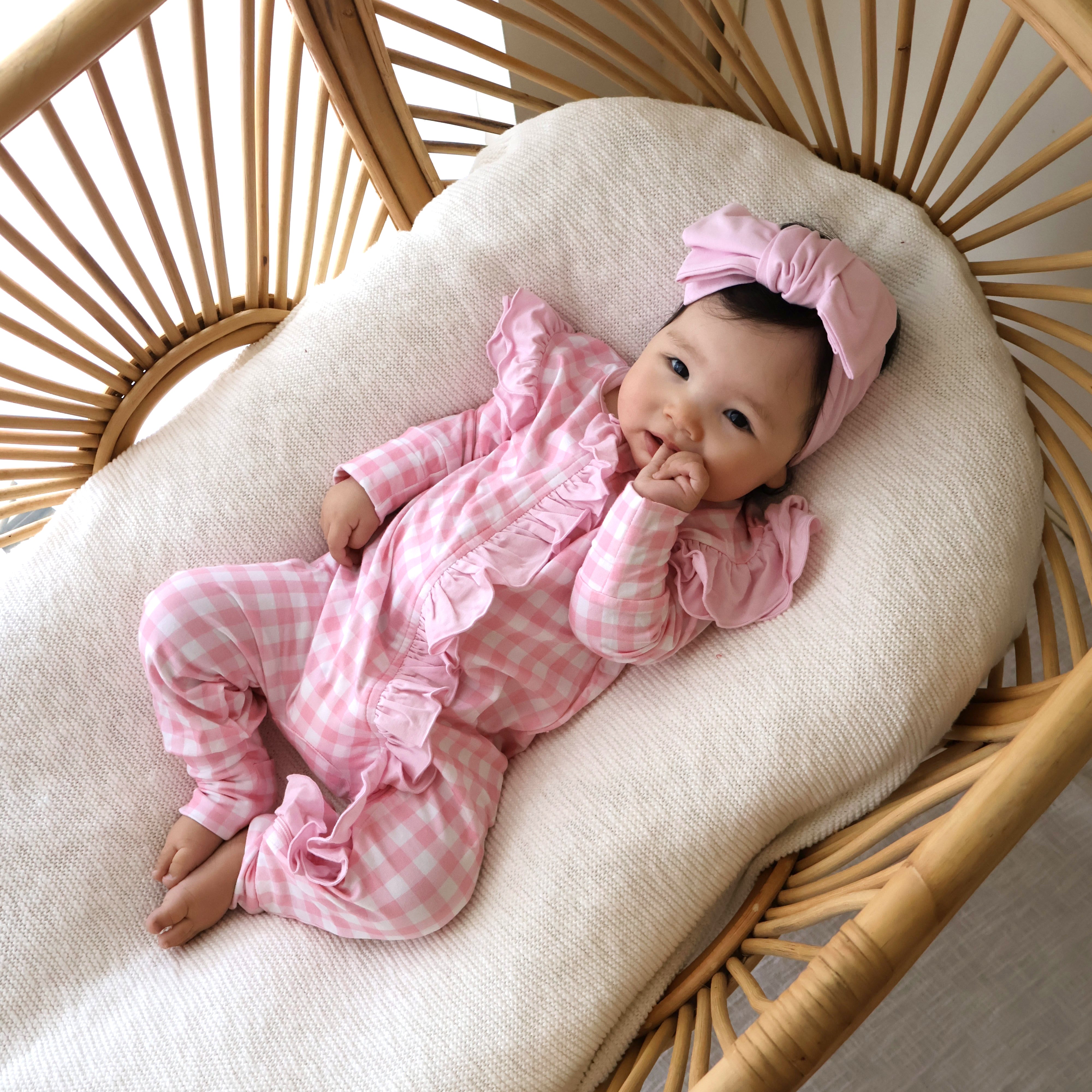 Baby in pink gingham frilly onesie outfit and dusky pink onesie bow headband lying on a woven chair with white cushion